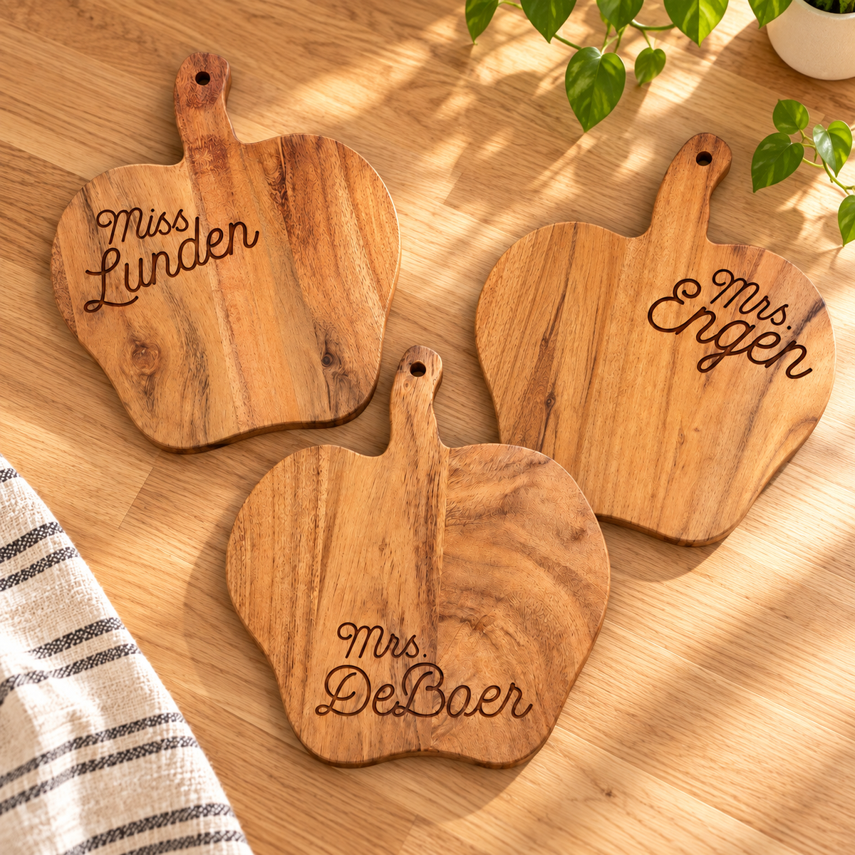 Three engraved apple-shaped teacher cutting boards lying flat on a wood kitchen counter